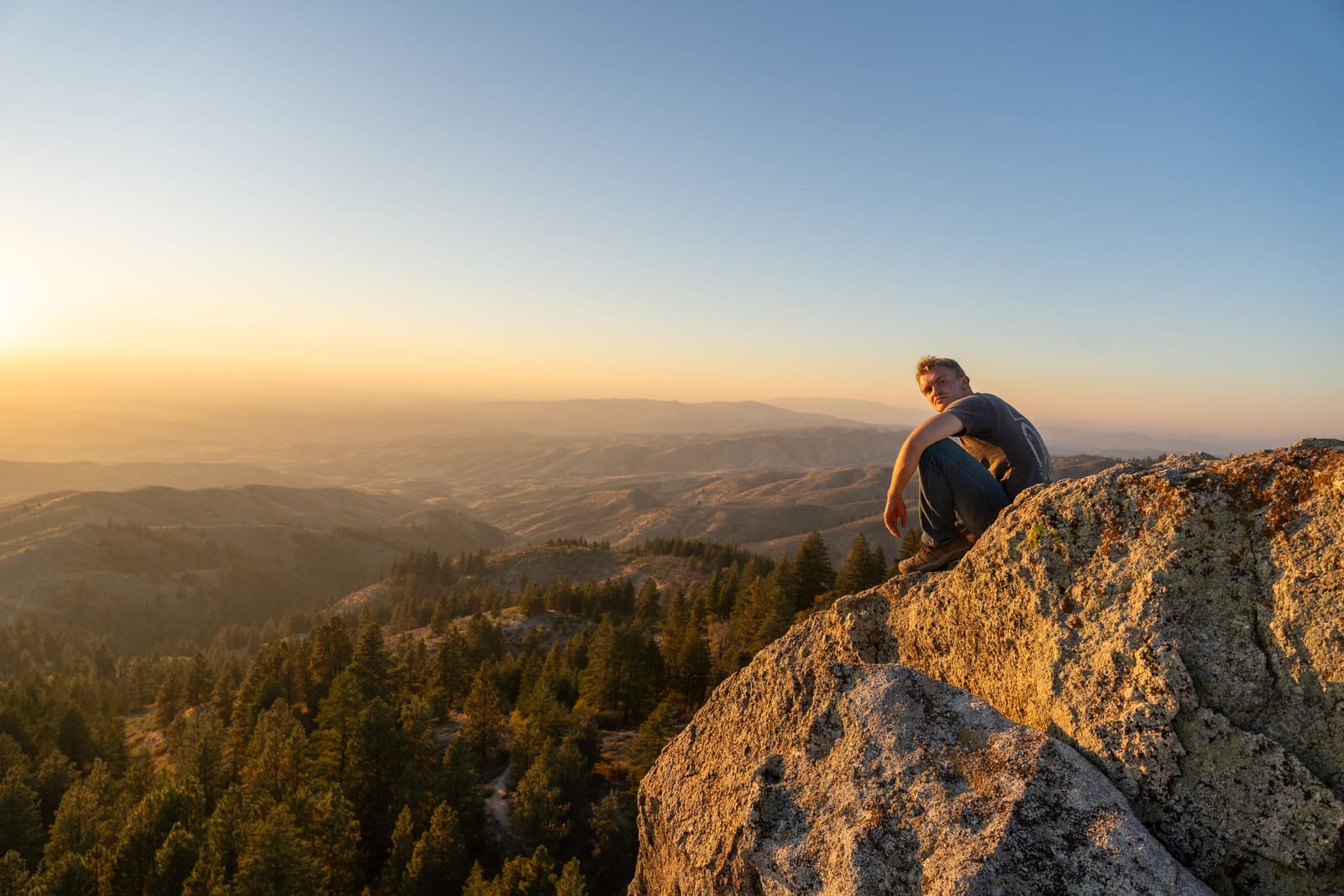 Figure seated on a rocky summit watching the sun set over forested Idaho ridges